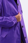 A person buttons a GINGHAM ULTRAVIOLET WINDBREAKER by The Windbreaker Jacket, featuring dark buttons, water resistance, elasticized cuffs, and a side pocket. Red nails contrast against a plain white background.