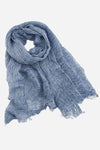 A light blue, textured Lightweight Linen Scarf made of ultra-breathable linen by looselyboho. The scarf has a slightly crinkled appearance and frayed edges, giving it an artisanal charm and relaxed look. It is draped and wrapped in a loose, flowing manner against a plain, light grey background.