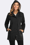 A woman is wearing a black hooded Elegant Plaid Raincoat Windbreaker from looselyboho made of high-quality, water-resistant fabric with her hands in the coat's pockets. She is smiling and standing against a plain white background. The lightweight raincoat has a front button closure and side pockets.