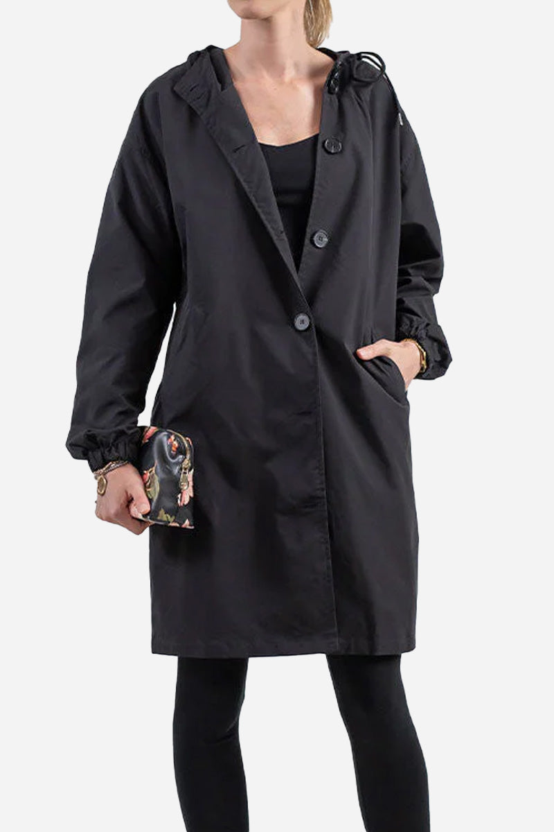 A person standing against a white background is wearing The WJacket's Water Resistant Oversized Hooded Windbreaker Rain Jacket with their left hand in their pocket. Showcasing timeless style and versatility, they are holding a small black clutch purse in their right hand and are dressed in black leggings and a black top.