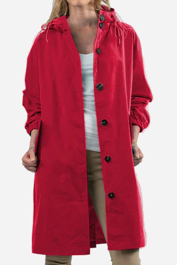 A person is wearing The WJacket’s Water Resistant Oversized Hooded Windbreaker Rain Jacket in red, embodying timeless style. The jacket features large black buttons and elasticated cuffs, adding to its versatility. They have a white top underneath and beige pants. The image focuses on the upper half of the body.