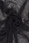 A close-up of a textured black scarf, artfully arranged in swirling, circular folds. The material appears delicate and slightly transparent, with varying layers creating a sense of depth and movement—perfect for all-weather use with the Color-POP Scarf by looselyboho.