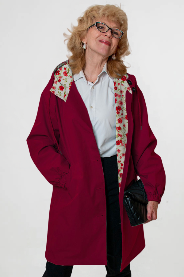A woman wearing glasses stands confidently in a looselyboho BURGUNDY BLOSSOM WINDBREAKER with a floral lining, paired with a white blouse and dark pants. Her black quilted handbag enhances her chic look, while the simple white background accentuates the elegance of her ensemble.