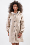 A person with curly hair wearing a BISCUIT BEIGE WINDBREAKER by looselyboho, a lightweight water-resistant raincoat with black buttons, stands against a plain white background. The beige coat, featuring an adjustable hood, has their right hand in one of the pockets while their left hand rests relaxed by their side.