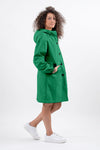 A person with curly hair stands in an EMERALD GREEN WINDBREAKER by The Windbreaker Jacket, featuring an adjustable hood and white sneakers. Their left hand is in a pocket, and they are slightly turned to the side, looking into the distance. The background is plain white.