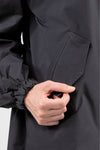 A close-up image of a person wearing a stylish looselyboho ONYX BLACK WINDBREAKER. The person's hand is shown pulling open a pocket on the lightweight water-resistant material. The jacket sleeve has an elastic band around the wrist. The background is plain white.